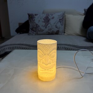 Peut inclure: Une lampe cylindrique de style tiki blanc cass&eacute; avec un motif de visage sculpt&eacute;. La lampe est &eacute;clair&eacute;e de l'int&eacute;rieur, projetant une lueur chaude. La lampe est sur une surface blanche, avec un cordon blanc.