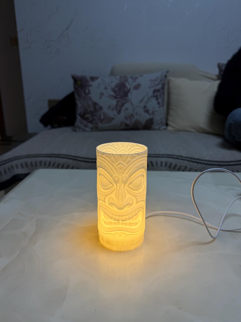 Peut inclure: Une lampe cylindrique de style tiki blanc cass&eacute; avec un motif de visage sculpt&eacute;. La lampe est &eacute;clair&eacute;e de l'int&eacute;rieur, projetant une lueur chaude. La lampe est sur une surface blanche, avec un cordon blanc.