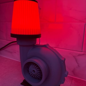 May include: A gray lamp shaped like a turbocharger with a red filter as the lampshade. The lamp sits on a black base and is lit with a red glow. The lamp is a unique decorative item.
