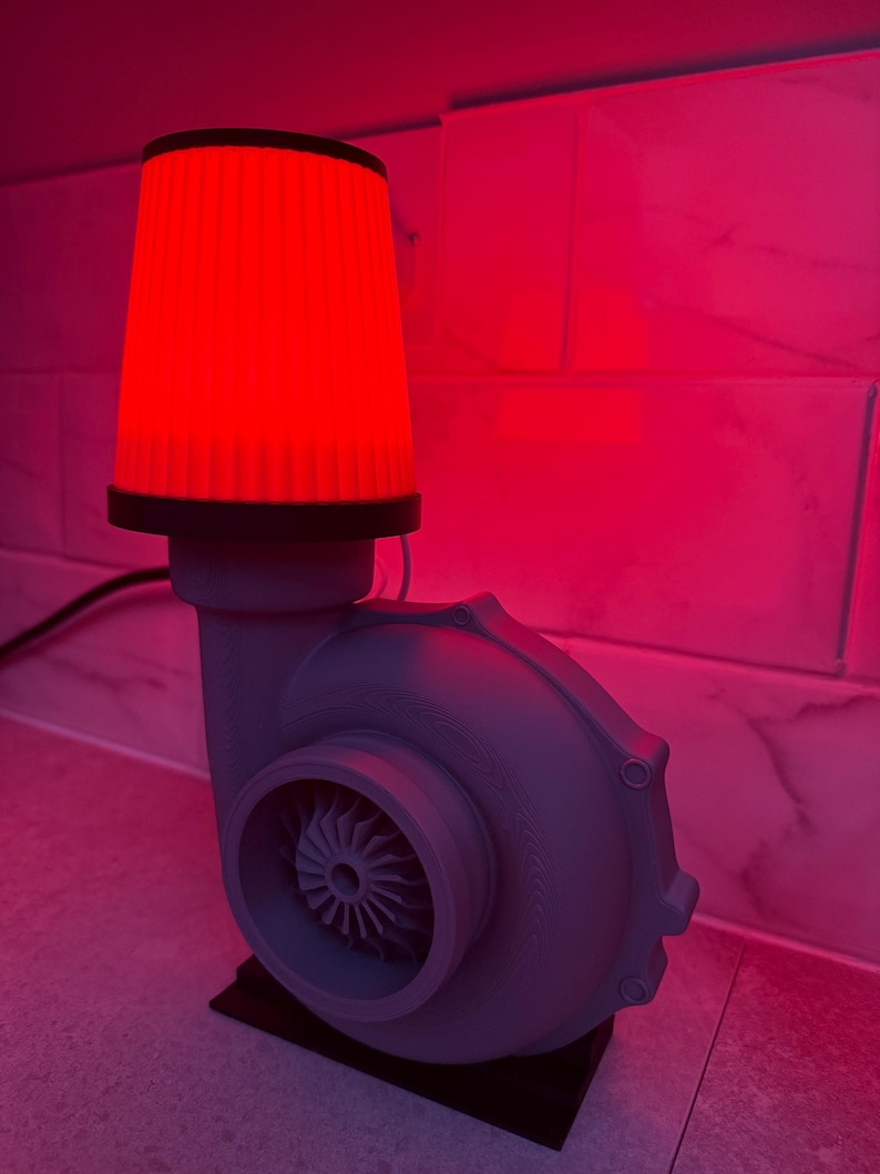 May include: A gray lamp shaped like a turbocharger with a red filter as the lampshade. The lamp sits on a black base and is lit with a red glow. The lamp is a unique decorative item.