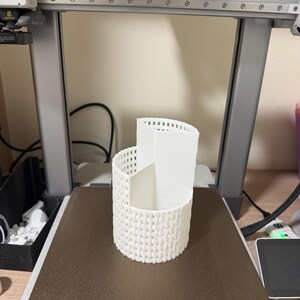 May include: A white, 3D-printed desk organizer with a woven basket design. The organizer has three compartments of varying heights and is sitting on a printer bed. The printer is in a workshop setting.