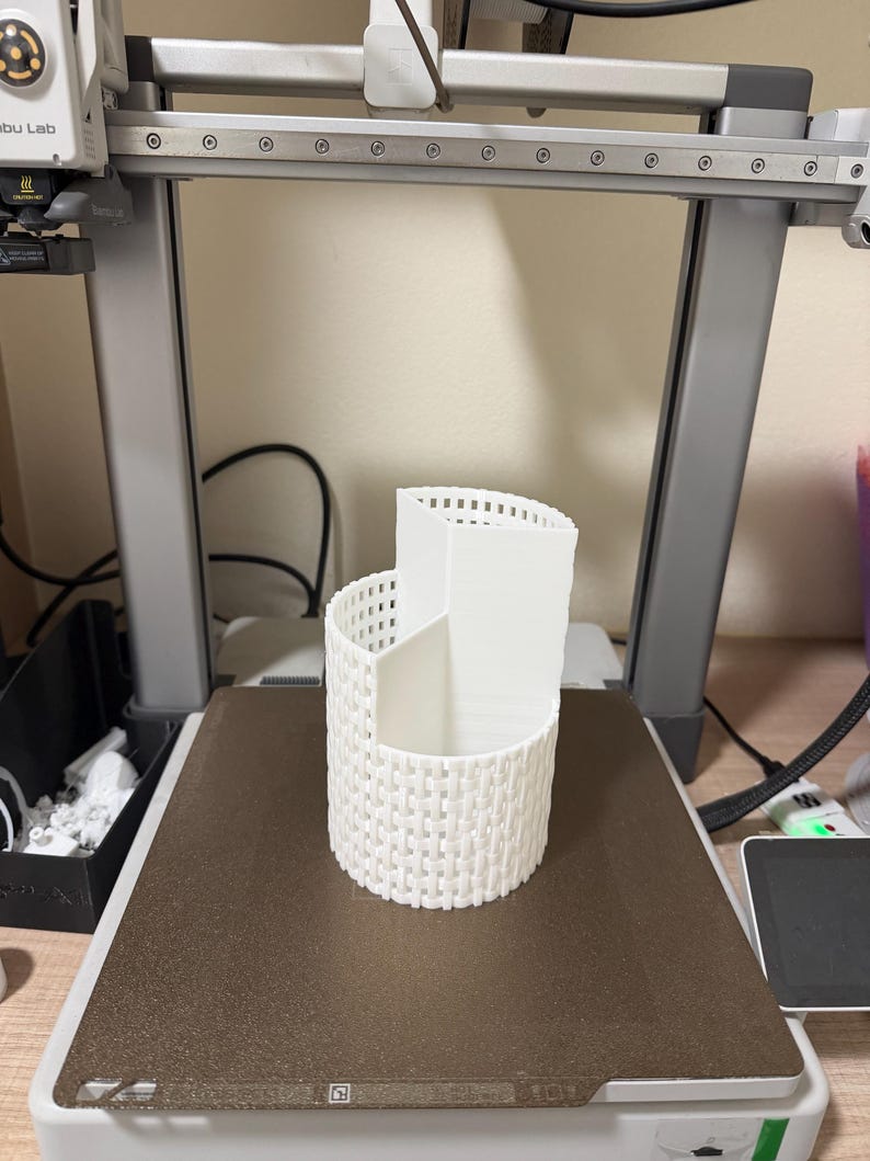 May include: A white, 3D-printed desk organizer with a woven basket design. The organizer has three compartments of varying heights and is sitting on a printer bed. The printer is in a workshop setting.