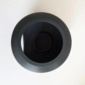 May include: A black, cylindrical object viewed from above, featuring a wide, textured rim. The interior is a deep black, with a smaller, circular base at the bottom. The object is set against a plain white background.