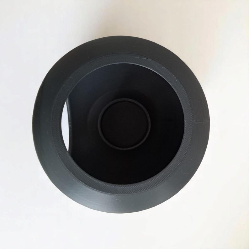 May include: A black, cylindrical object viewed from above, featuring a wide, textured rim. The interior is a deep black, with a smaller, circular base at the bottom. The object is set against a plain white background.