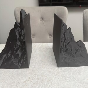 May include: A pair of black mountain-shaped bookends. Each bookend has a rectangular base and a detailed mountain range design on the front. The bookends are made of a solid, dark material and are designed to hold books upright.