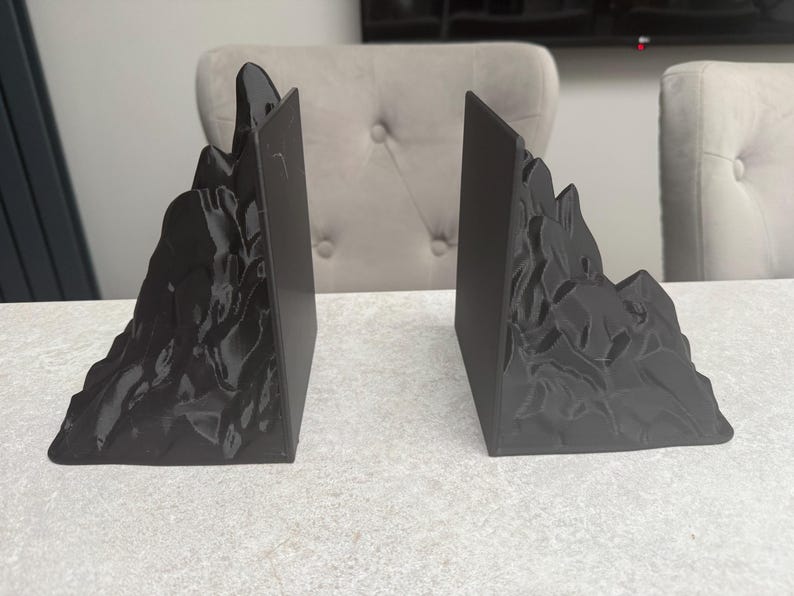 May include: A pair of black mountain-shaped bookends. Each bookend has a rectangular base and a detailed mountain range design on the front. The bookends are made of a solid, dark material and are designed to hold books upright.
