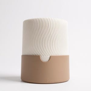 May include: A cylindrical ceramic container with a two-tone design. The upper portion is off-white with a wavy, ridged texture, while the lower section is a solid tan color. The container has a small, curved opening on the front.