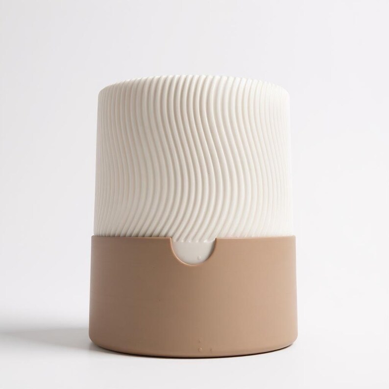May include: A cylindrical ceramic container with a two-tone design. The upper portion is off-white with a wavy, ridged texture, while the lower section is a solid tan color. The container has a small, curved opening on the front.