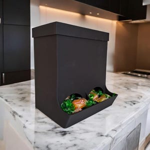 May include: A black candy dispenser with a rectangular shape and a small tray at the bottom. The dispenser is filled with wrapped candies, visible through two circular openings. The dispenser sits on a marble countertop.