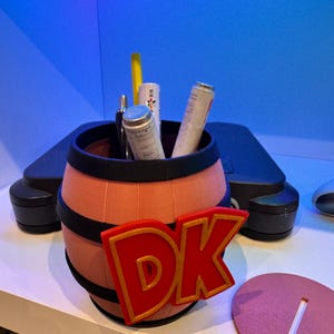 May include: A barrel-shaped container with the letters "DK" in red and gold. The barrel is peach-colored with black bands and holds several cylindrical items. A pink oval base with a slot is next to the barrel.