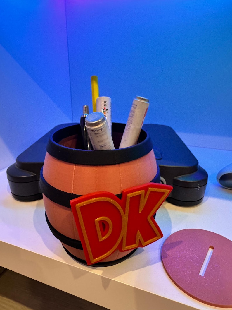 May include: A barrel-shaped container with the letters "DK" in red and gold. The barrel is peach-colored with black bands and holds several cylindrical items. A pink oval base with a slot is next to the barrel.