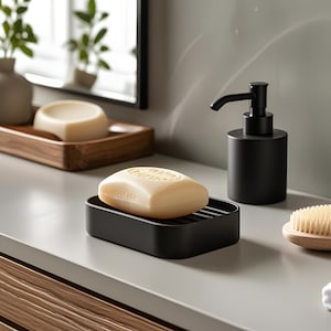 May include: A light beige bar of soap rests on a black soap dish. A black soap dispenser and a wooden tray with soap are also visible. The scene is set on a light gray countertop, suggesting a bathroom setting.