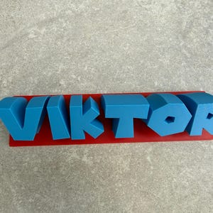 Op de afbeelding: Een blauwe 3D-geprinte naam, "VIKTOR", wordt weergegeven op een rode rechthoekige basis. De letters zijn in een vet, blokkerig lettertype en de naam is gecentreerd op de basis. De achtergrond is een lichtgrijze ondergrond.