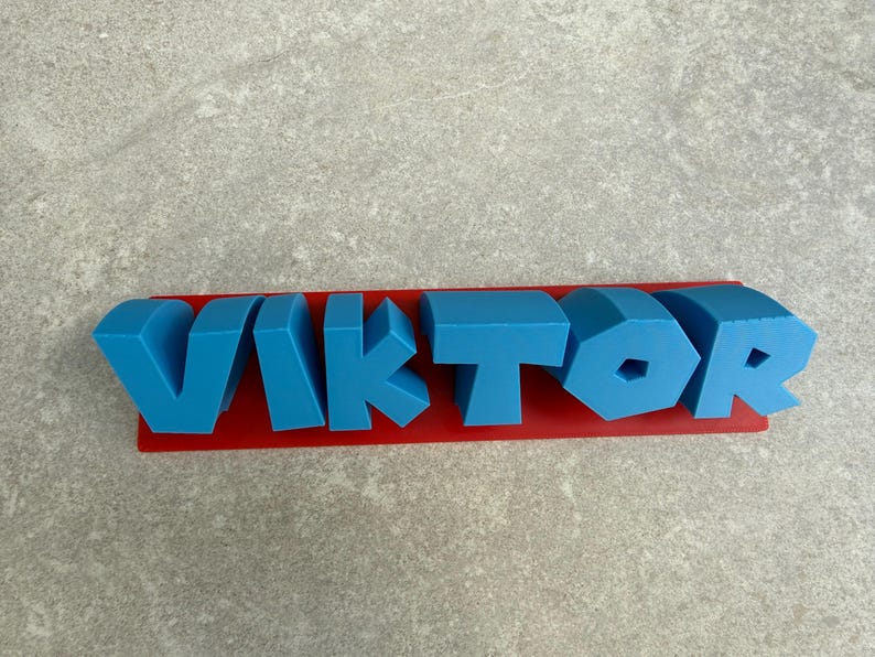 Op de afbeelding: Een blauwe 3D-geprinte naam, "VIKTOR", wordt weergegeven op een rode rechthoekige basis. De letters zijn in een vet, blokkerig lettertype en de naam is gecentreerd op de basis. De achtergrond is een lichtgrijze ondergrond.