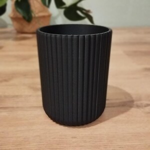 May include: A black cylindrical planter with vertical ribbed detailing. The planter is made of a matte material and sits on a wooden surface. A plant in a woven basket is visible in the background.