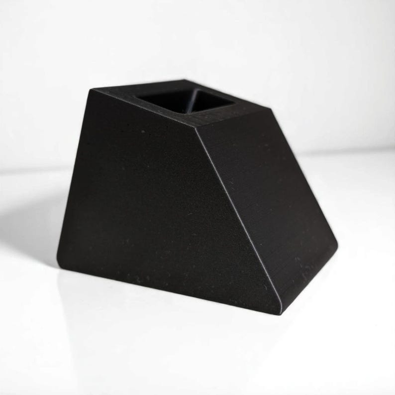 May include: A black, geometric candle holder with a trapezoidal shape. The top features two square openings for candles. The holder is made of a solid, matte black material and sits on a white surface.