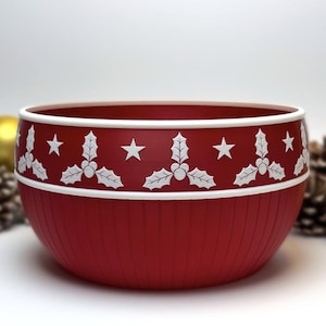 May include: A red bowl with a white rim and a decorative band featuring white holly leaves and stars. The bowl has vertical lines and is likely for serving or display. Festive holiday decor.