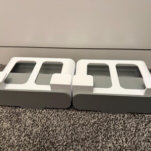 May include: Two white and gray organizers, each with a rectangular base and a top section featuring two rectangular openings. The organizers have a car-like shape, with a gray base and white upper structure. They sit on a textured, gray carpet.
