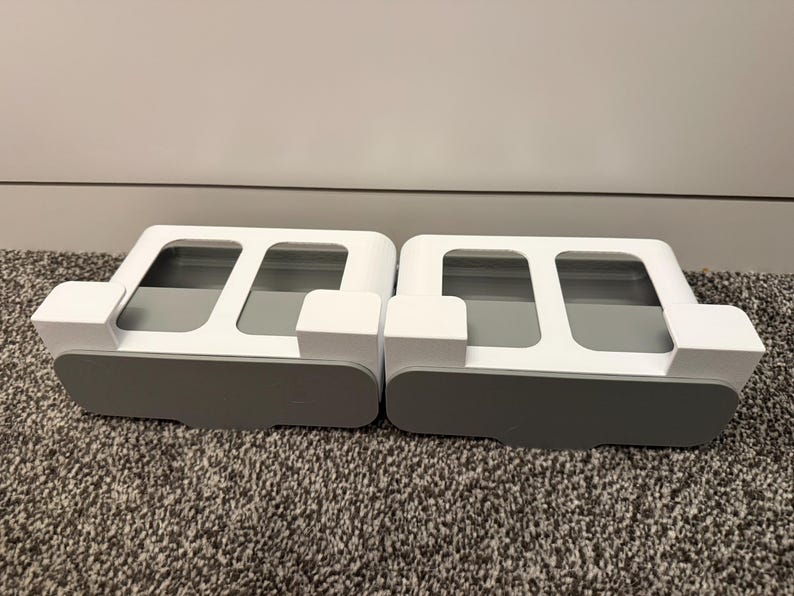 May include: Two white and gray organizers, each with a rectangular base and a top section featuring two rectangular openings. The organizers have a car-like shape, with a gray base and white upper structure. They sit on a textured, gray carpet.