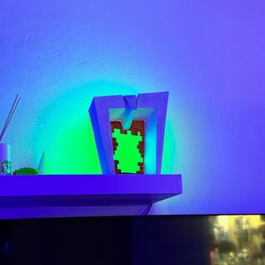 May include: A modern, geometric wooden sculpture with a green pixelated design in the center. The sculpture is illuminated by a vibrant green and blue light, creating a striking visual effect. The piece is displayed on a white shelf against a blue wall.
