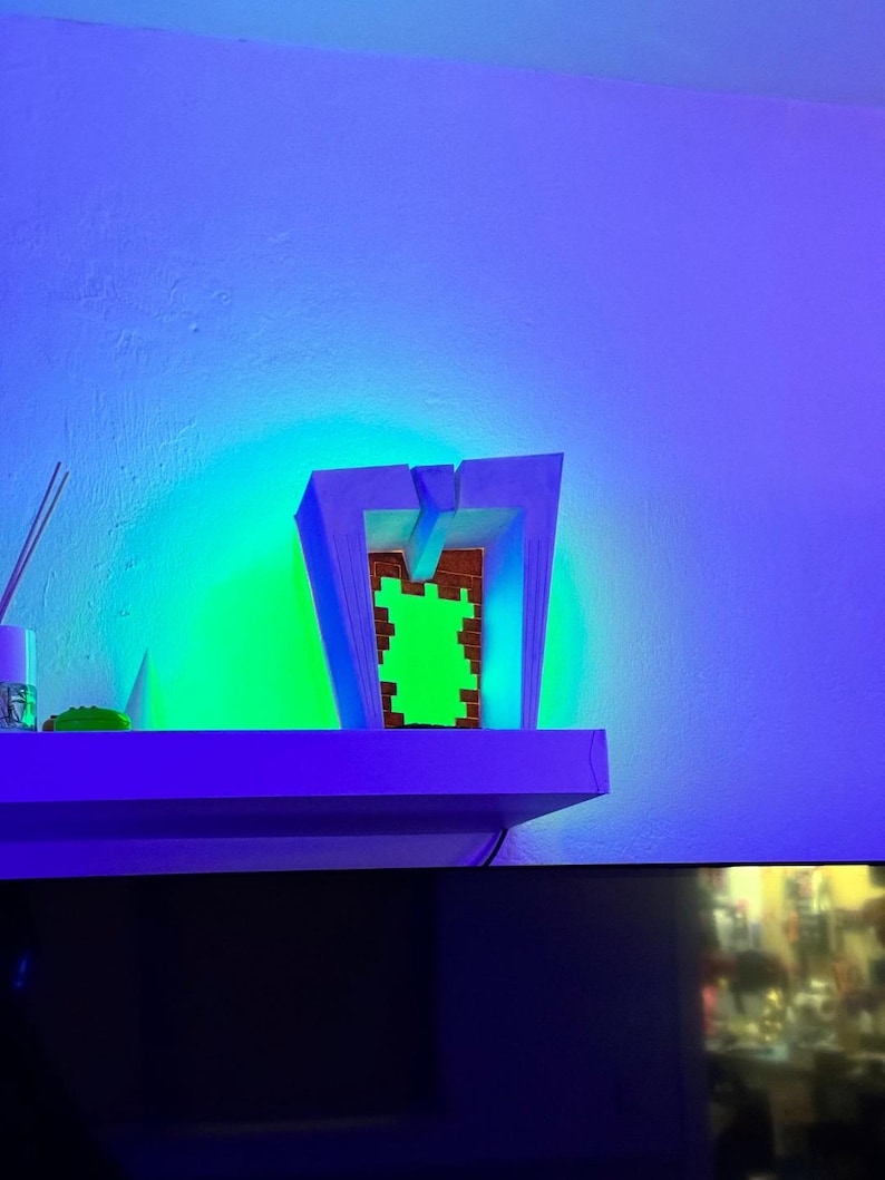 May include: A modern, geometric wooden sculpture with a green pixelated design in the center. The sculpture is illuminated by a vibrant green and blue light, creating a striking visual effect. The piece is displayed on a white shelf against a blue wall.