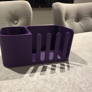 May include: A solid purple utensil holder with a divided compartment and a slotted side. The holder is made of a matte material and sits on a light gray surface. The background includes two upholstered chairs.