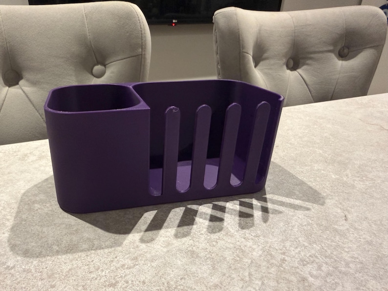 May include: A solid purple utensil holder with a divided compartment and a slotted side. The holder is made of a matte material and sits on a light gray surface. The background includes two upholstered chairs.