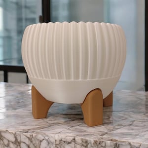 Pode incluir: Um vaso branco e canelado com formato arredondado, sustentado por tr&ecirc;s pernas marrons. O vaso tem uma borda ondulada e fica sobre uma superf&iacute;cie de m&aacute;rmore. Uma pe&ccedil;a decorativa moderna para plantas de interior.