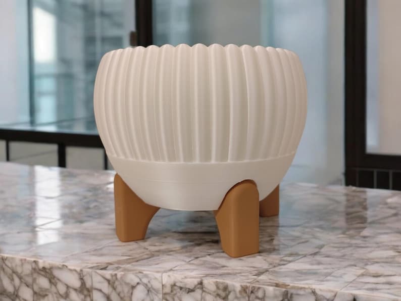 Pode incluir: Um vaso branco e canelado com formato arredondado, sustentado por tr&ecirc;s pernas marrons. O vaso tem uma borda ondulada e fica sobre uma superf&iacute;cie de m&aacute;rmore. Uma pe&ccedil;a decorativa moderna para plantas de interior.