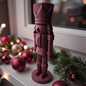May include: A burgundy-colored nutcracker figurine stands on a white windowsill, surrounded by Christmas ornaments and greenery. The figurine has a detailed design, wearing a crown and uniform. Decorative Christmas balls and lights add to the festive scene.
