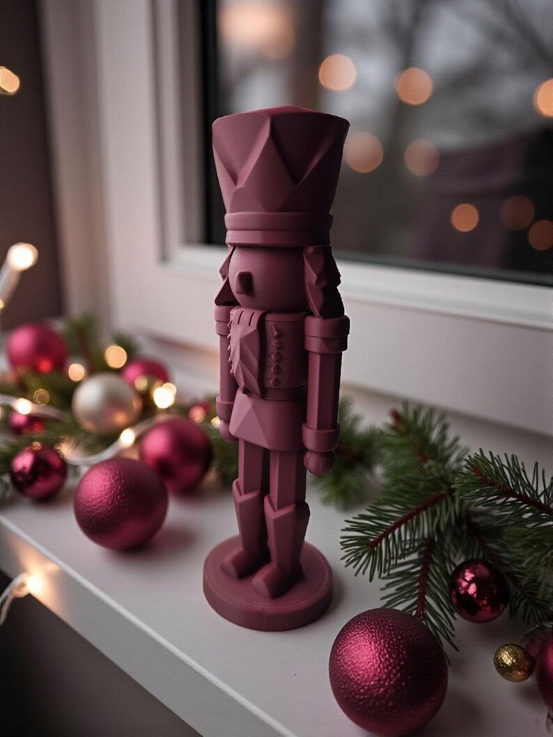 May include: A burgundy-colored nutcracker figurine stands on a white windowsill, surrounded by Christmas ornaments and greenery. The figurine has a detailed design, wearing a crown and uniform. Decorative Christmas balls and lights add to the festive scene.