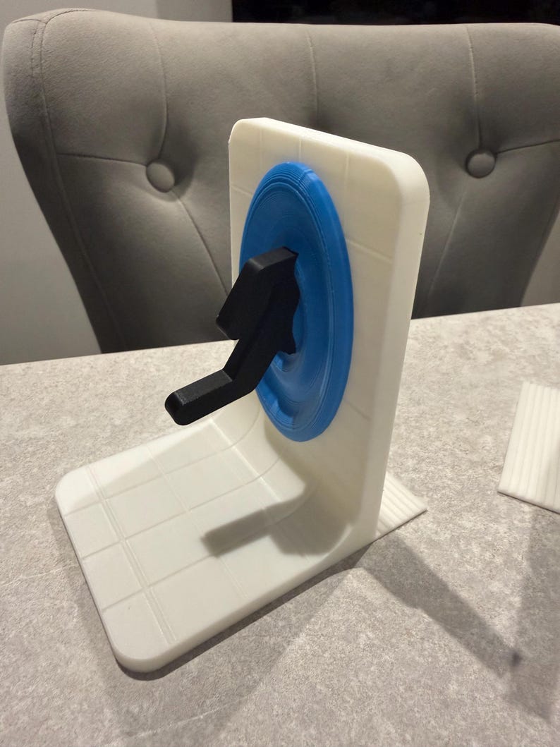 May include: A white and blue 3D-printed bookend featuring a black arrow design. The bookend has a square shape with a blue circle and a black arrow. The base is white with a grid pattern.