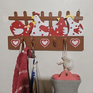 Puede incluir: Un perchero de pared de madera marr&oacute;n con cuatro figuras de gallinas estilizadas en rojo y blanco, con un dise&ntilde;o de valla marr&oacute;n en la parte superior. Debajo hay cuatro ganchos y detalles en forma de coraz&oacute;n. Varios objetos cuelgan de los ganchos, incluyendo una toalla roja.