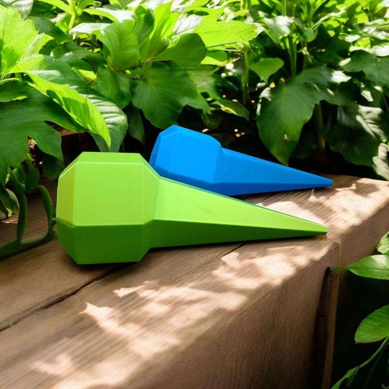 May include: Two plastic plant watering spikes, one green and one blue, rest on a wooden surface. The spikes have a geometric design and are set against a backdrop of green foliage. These are designed to slowly release water into the soil.