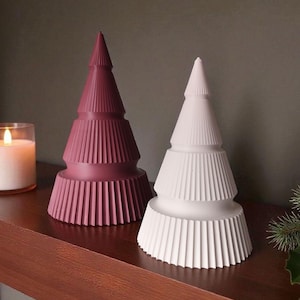 May include: Two decorative Christmas tree figurines. One is a deep rose color, the other is white. Both have a tiered design with vertical ribbed detailing. They are displayed on a wooden surface.