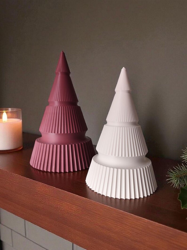 May include: Two decorative Christmas tree figurines. One is a deep rose color, the other is white. Both have a tiered design with vertical ribbed detailing. They are displayed on a wooden surface.