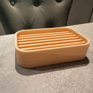 May include: A rectangular, peach-colored soap dish with a slatted top. The dish is made of a solid material and sits on a gray surface. The design is simple and modern, suitable for a bathroom or kitchen.
