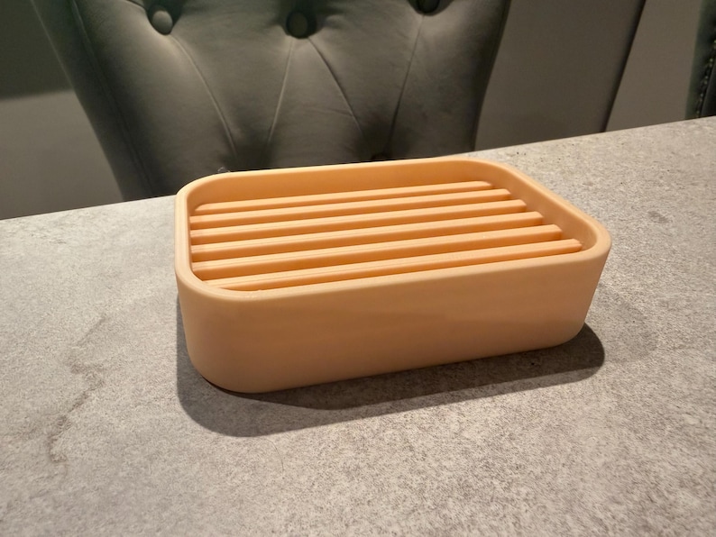 May include: A rectangular, peach-colored soap dish with a slatted top. The dish is made of a solid material and sits on a gray surface. The design is simple and modern, suitable for a bathroom or kitchen.