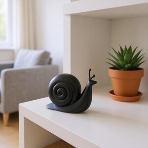 May include: A matte black snail figurine with a spiraled shell sits on a white surface. The snail has two antennae and is positioned next to a small potted succulent in a terracotta pot, creating a decorative display.