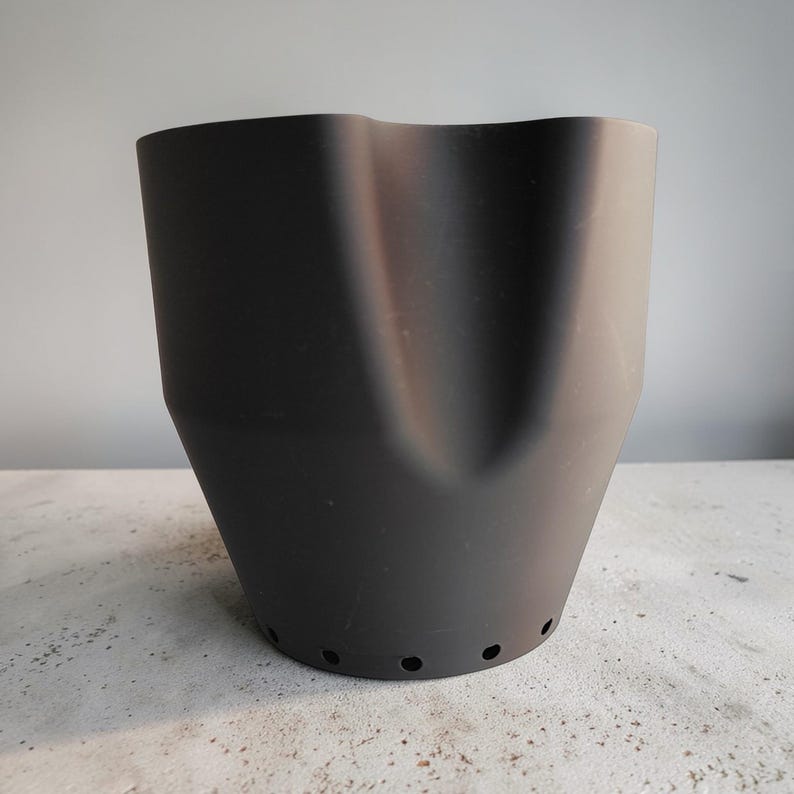 May include: A matte black planter with a unique, curved design. The planter has a wide top that narrows towards the base, with several small drainage holes. The overall shape is reminiscent of a stylized vase or pot, suitable for indoor or outdoor use.