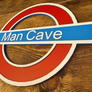 May include: A sign with a red circle and a blue rectangle, featuring the text "Man Cave" in white. The sign has a white border and is set against a wooden background.
