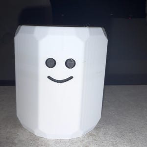 May include: A white, geometric-shaped container with a smiling face design. The container has a polygonal shape and a flat top. The face features two black, circular eyes and a curved, black smile. The object is likely a decorative item or a small storage container.
