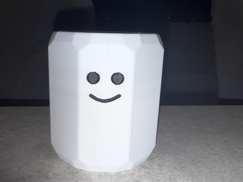 May include: A white, geometric-shaped container with a smiling face design. The container has a polygonal shape and a flat top. The face features two black, circular eyes and a curved, black smile. The object is likely a decorative item or a small storage container.