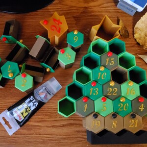 May include: An Advent calendar in the shape of a Christmas tree, with green and brown hexagonal compartments numbered 1-24. Additional numbered hexagonal pieces and star-shaped pieces are scattered around. A tube of glue is also present.
