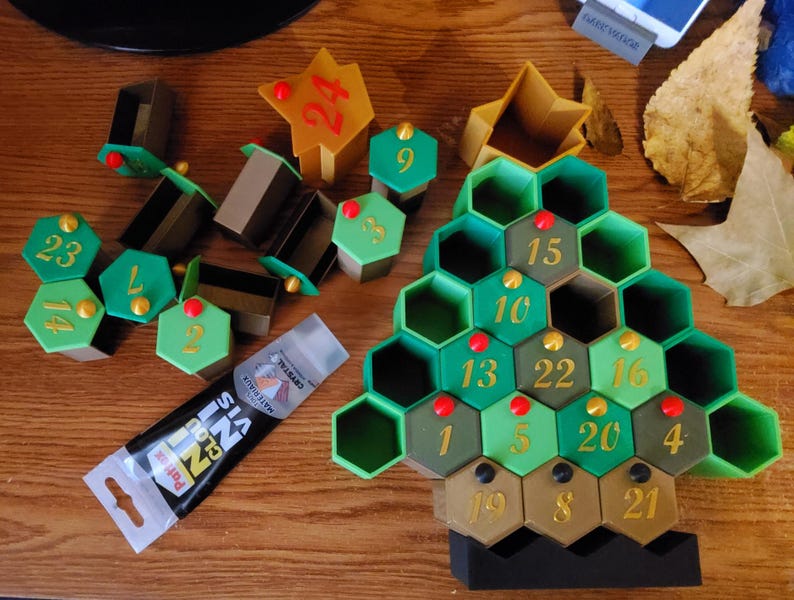 May include: An Advent calendar in the shape of a Christmas tree, with green and brown hexagonal compartments numbered 1-24. Additional numbered hexagonal pieces and star-shaped pieces are scattered around. A tube of glue is also present.