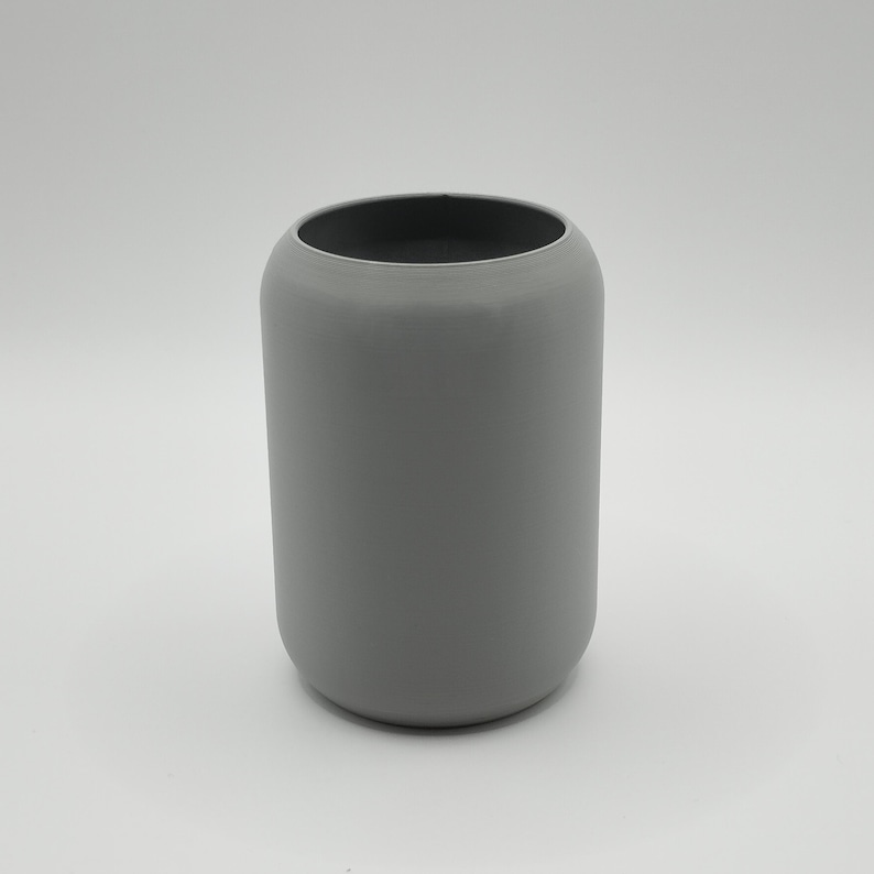 May include: A cylindrical, gray ceramic vase with a rounded top and a dark interior. The vase has a smooth, matte finish and a simple, modern design. The vase is suitable for holding flowers or as a decorative item.