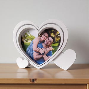 May include: A white heart-shaped photo frame displaying a photograph of a couple. The frame features a layered design and two small heart accents. The frame is on a wooden surface.