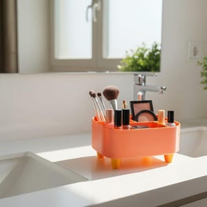 May include: An orange makeup organizer with yellow feet, holding various makeup brushes, lipsticks, and compacts. The organizer sits on a white countertop in a bathroom setting, with a mirror and sink in the background.