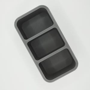 May include: A gray, rectangular organizer with three compartments. Each compartment has a textured, dark gray interior. The organizer is made of a solid material and is designed to hold small items.