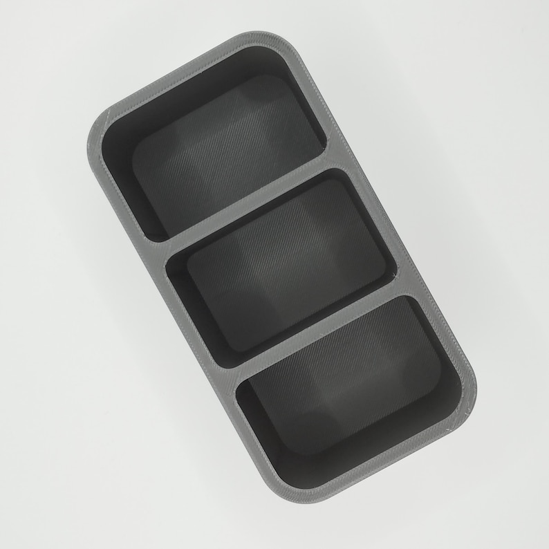 May include: A gray, rectangular organizer with three compartments. Each compartment has a textured, dark gray interior. The organizer is made of a solid material and is designed to hold small items.